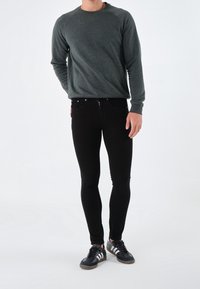 Man som bär en mörkgrå långärmad tröja, svarta tajta jeans och svarta sneakers med vita ränder, stående mot en vit bakgrund.