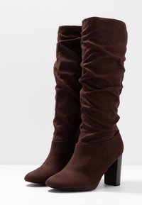 Bruna mockaimocka knähöga stövlar med en samlad design, spetsiga tår och en kraftig klack, med en slät textur och utan synlig hårdvara.