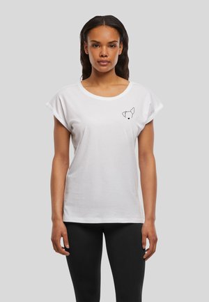 Femme portant un t-shirt blanc avec un petit contour noir d'un visage de chien sur le côté gauche de la poitrine et un pantalon noir, debout devant un fond gris.