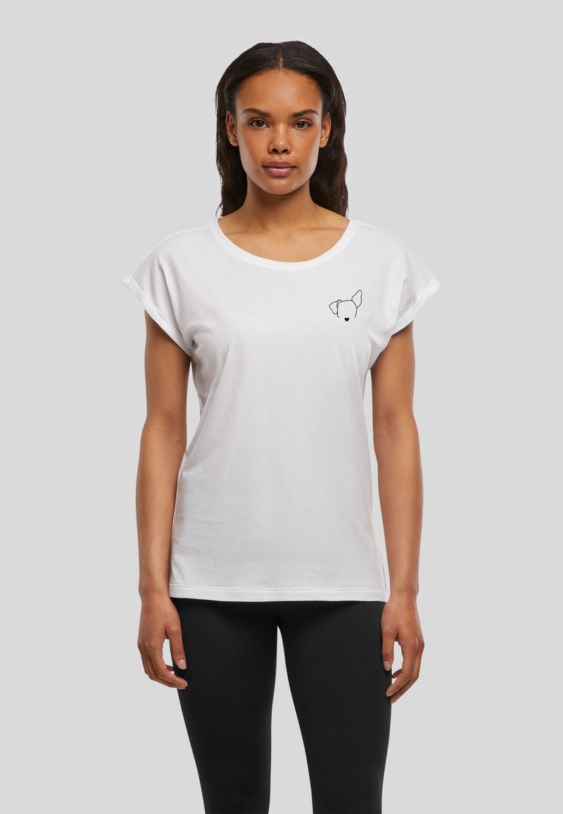 Femme portant un t-shirt blanc avec un petit contour noir d'un visage de chien sur le côté gauche de la poitrine et un pantalon noir, debout devant un fond gris.