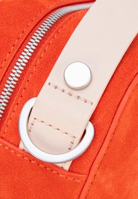 Detail van een oranje suède tas met een crèmekleurige leren band, voorzien van een zilverkleurige metalen ring en drukknop, waarbij de rits langs de rand wordt benadrukt.
