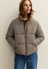 Bruine houndstooth-gepatroneerde pufferjas met een hoge kraag, ritssluiting aan de voorkant en zijzakken, gedragen met een bruine trui en jeans.