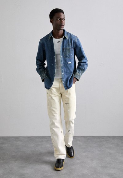 Chemise en denim avec poche applique, bleu clair ; t-shirt blanc en dessous, pantalon crème déchiré et chaussures noires avec semelles beige.