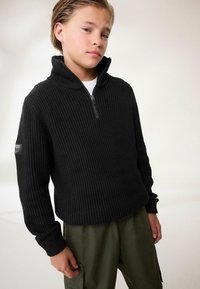 Pullover nero a coste con colletto alto e zip a quarto, abbinato a pantaloni cargo verdi. Presenta una patch con logo sulla manica.