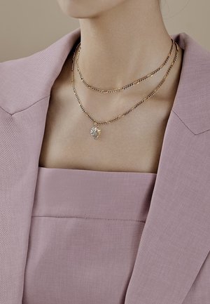 Colliers en chaîne dorée à deux rangs avec un pendentif feuille, portés sur un haut mauve à col carré et un blazer assorti.