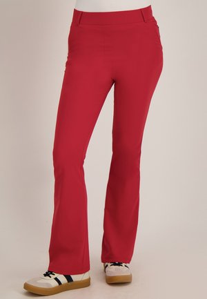 Rode slim-fit broek gedragen met witte sneakers met zwarte strepen en gumzolen, getoond op een neutrale achtergrond.