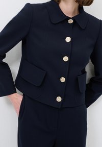Kvinna i marinblå skräddarsydd jacka med texturerade guldknappar och matchande högmidjade byxor, med handen i fickan, mot vit bakgrund.