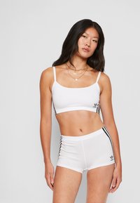 Conjunto de top curto branco e calções, apresentando alças finas, uma textura suave e riscas laterais pretas com um detalhe de logótipo à esquerda.