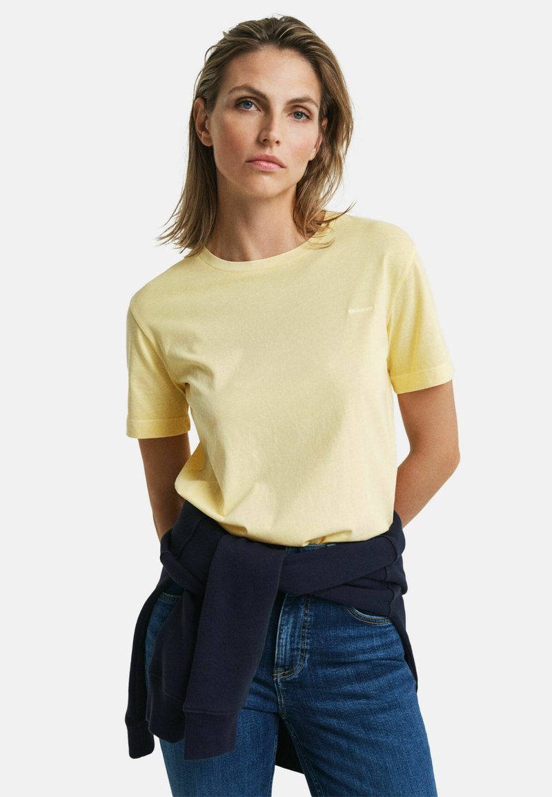 Lichtgele katoenen T-shirt met korte mouwen en een ronde hals. Een donkere trui is om de taille geknoopt, gecombineerd met blauwe spijkerbroeken.