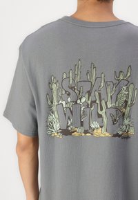 Szara bawełniana koszulka z dużą grafiką z napisem "STAY WILD" otoczoną kaktusami i roślinami w zielonych i brązowych odcieniach.