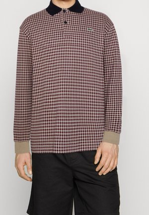 Homme portant un polo à manches longues à motif pied-de-poule bordeaux et beige avec col noir et poignets beiges, associé à un short noir.