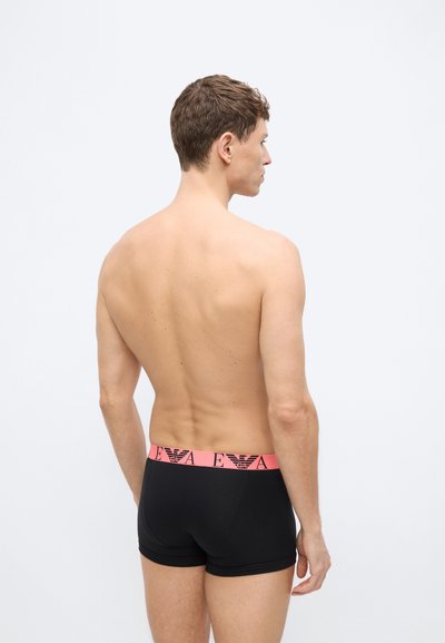 Άνδρας, φαίνεται από πίσω, φοράει μαύρα μποξεράκια με ροζ ζώνη μέσης που φέρει επαναλαμβανόμενο μαύρο λογότυπο και γράμματα.