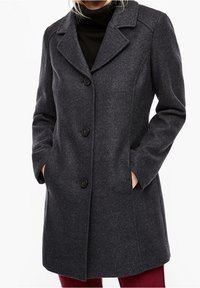 Donna che indossa un cappotto di lana grigio scuro con tre bottoni e tasche laterali, abbinato a un dolcevita nero e pantaloni rossi.