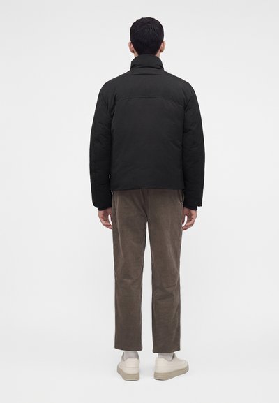 Homme debout de dos portant une veste noire, un pantalon marron, des chaussettes blanches et des baskets beiges sur un fond blanc uni.