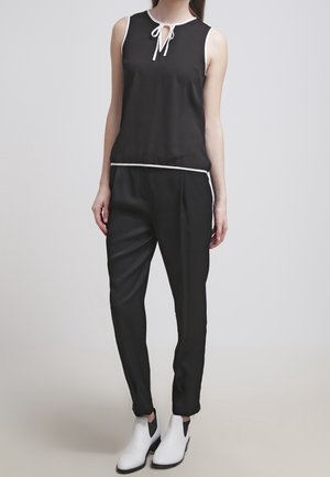 Top senza maniche nero con un fiocco davanti e un accento bianco, abbinato a pantaloni neri a sigaretta e stivaletti bianchi, su uno sfondo neutro.