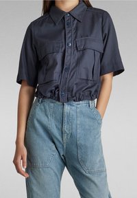 Marinblus med kort ärm och två framfickor, knäppning och rynkad nederkant. Matchad med ljusblå jeans med hög midja.