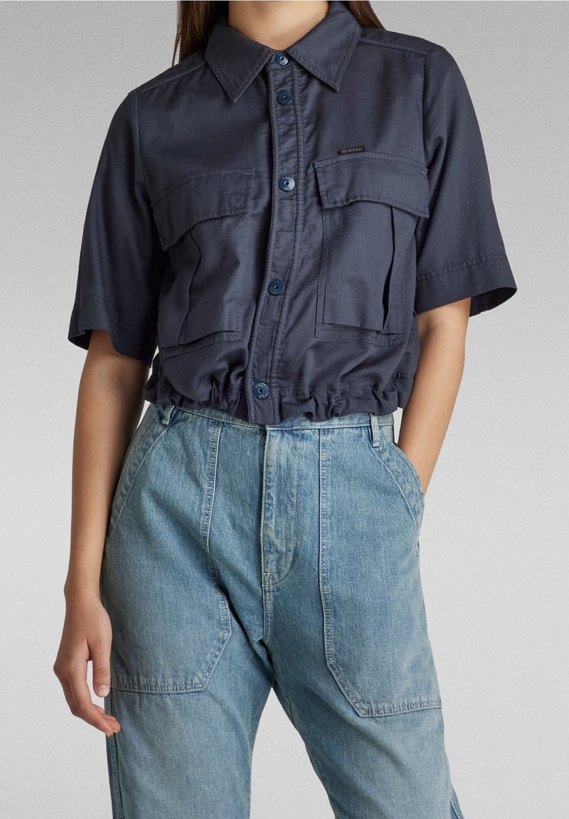 Marinblus med kort ärm och två framfickor, knäppning och rynkad nederkant. Matchad med ljusblå jeans med hög midja.