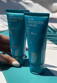 Två turkosa tuber av KIKO Solskyddskräm, SPF 80 och SPF 50, med vit text och guldaccenter, placerade på en blå bakgrund.