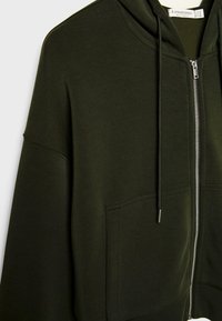 Dark green zip-up hoodie with front pockets, drawstring hood, and visible metal zipper, showing upper torso and sleeve area.