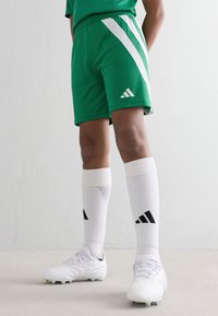 Person wearing green soccer shorts, white knee-high socks with black Adidas logo, and white cleats standing against a white wall.