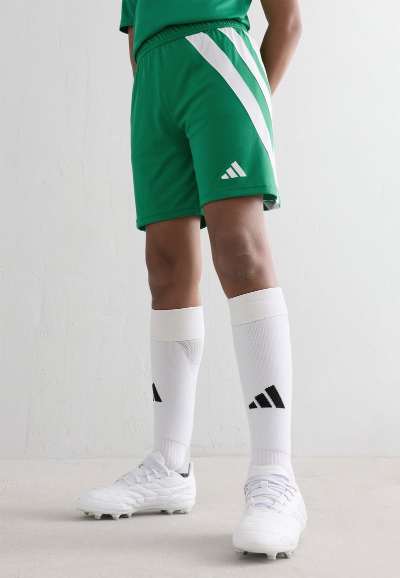 Person wearing green soccer shorts, white knee-high socks with black Adidas logo, and white cleats standing against a white wall.