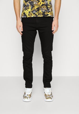 Strakzittende zwarte broek gecombineerd met witte sneakers met goudkleurige en zwarte barokmotieven, gedragen door een persoon met ontspannen handen langs het lichaam.