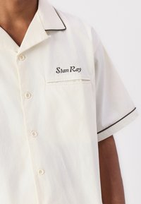 Cream-colored short-sleeve shirt with a pocket, black piping along the collar and sleeves, featuring embroidered text “Stan Ray.”