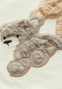 Bestickter Plüschbär auf einem cremefarbenen Stoff; verfügt über einen braunen Felltextur, schwarze Augen, gestickte Details und runde Musterakzente.