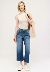 Haut sans manches beige avec col montant, associé à un jean bleu cropped à délavage léger. Des baskets blanches complètent le look. Un cardigan marron jeté sur le bras.