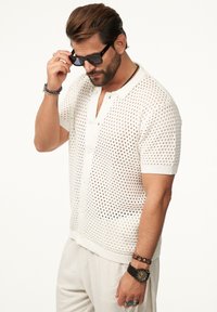 Weißes, gestricktes Polohemd mit kurzen Ärmeln und offenem Netzdesign, Knopfleiste und kontrastierendem Kragenbesatz. Person trägt Sonnenbrillen und Schmuck.