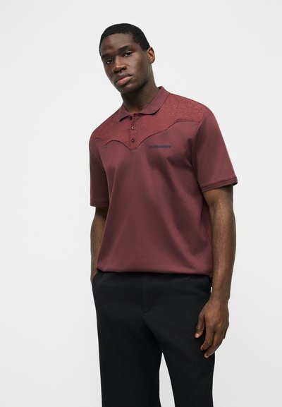 Νεαρός άνδρας που φοράει μαροον κοντομάνικη πόλο μπλούζα με μαύρο παντελόνι, στέκεται μπροστά σε ένα απλό ανοιχτό φόντο.