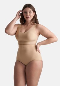 Kvinna med medellångt brunt hår som bär en beige sömlös bh-top och högmidjade underkläder, posera med en hand på höften och en hand som rör vid håret.