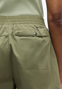 Κοντινό πλάνο σορτς με ελαστική μέση σε ελιά πράσινο χρώμα, με τσέπη στο πίσω μέρος που κλείνει με κουμπί και μερικώς ορατό χέρι.