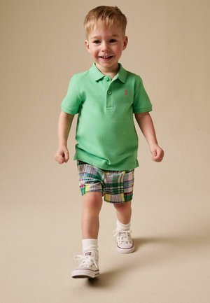 Smilende småbarn går fremad iført en grøn poloshirt, ternede shorts, hvide sokker og hvide sneakers på en ensartet beige baggrund.