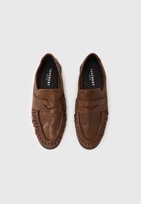 Καφέ δερμάτινα μοκασίνια με λείο φινίρισμα, διακοσμητικές ραφές κατά μήκος των πλευρών και μαύρη επίπεδη σόλα. Ονομασία εμπορικού σήματος στο εσωτερικό του παπουτσιού.