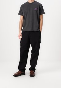 Grå bomullströja med korta ärmar och lila grafisk tryck på vänster bröst, tillsammans med svarta cargobyxor och bruna loafers.