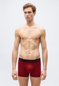 Νέος άνδρας με σγουρά μαλλιά που φοράει κόκκινα εσώρουχα μποξέρ της Nike,  στέκεται μπροστά σε απλό ανοιχτό γκρι φόντο, κοιτώντας ελαφρώς προς τα δεξιά.