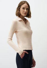 Lichtroze polo met lange mouwen, voorzien van een textuur in wafelstructuur en een contrasterende kraag. Relaxte pasvorm, getoond met een donkere broek.