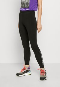 Leggings noirs au design ajusté, arborant un logo blanc sur la jambe gauche, associés à des chaussures de sport multicolores.