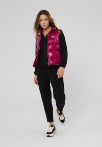 Magenta, glanzend puffer vest met een hoge kraag, gedragen over een zwart top en broek. De outfit is gecombineerd met zwart en gouden sneakers.