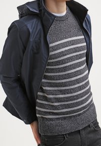 Persona che indossa un maglione a righe grigie e bianche sotto un giubbotto blu non zipato, in piedi con una mano in tasca su uno sfondo neutro.