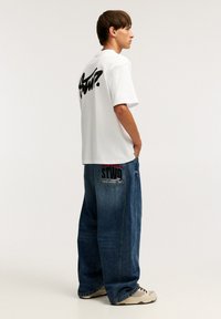 Hvid t-shirt med en sort grafik på ryggen, parret med løse blå denim jeans med et rødt og sort patch på lommen.