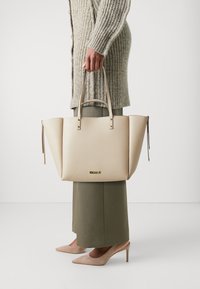 Beige strukturovaná taška s dvěma horními uchy, texturovaným povrchem, dekoračními komponenty a bočními zipy. Nosit s pleteným kardiganem a kalhotami.