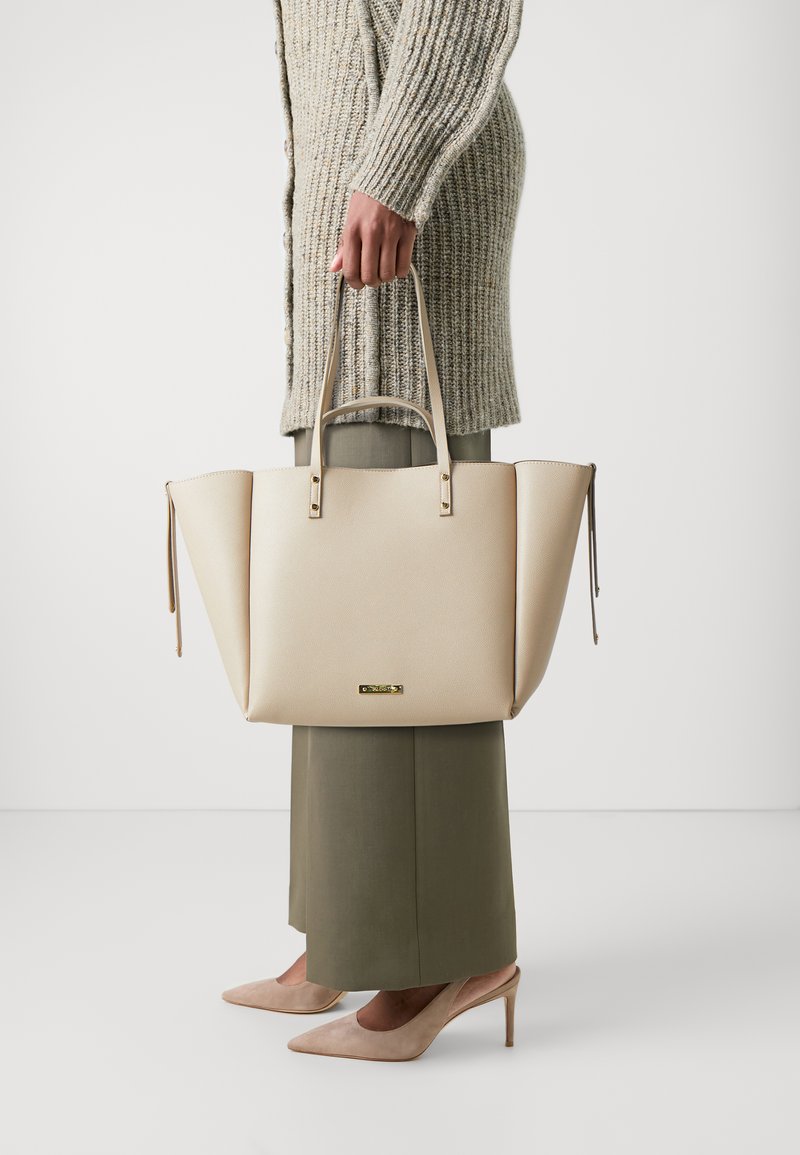 Beige strukturovaná taška s dvěma horními uchy, texturovaným povrchem, dekoračními komponenty a bočními zipy. Nosit s pleteným kardiganem a kalhotami.