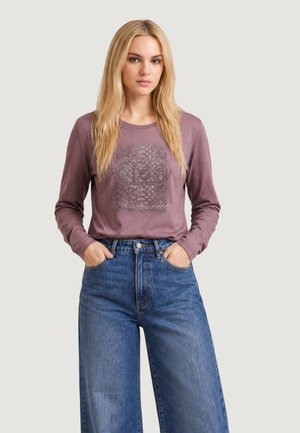 Joven mujer con pelo largo y rubio que lleva una camisa de manga larga color malva con un patrón gráfico y vaqueros azules de cintura alta con las manos en los bolsillos.