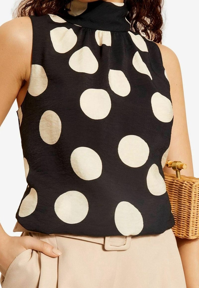Femme portant un haut noir sans manches à gros pois blancs et un pantalon beige taille haute, tenant un sac à main tressé.