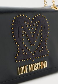 Melnas ādas rokassoma ar izvirzītu sirds dizainu tumšā audumā, rotāta ar zeltītiem kniedēm un zelta "LOVE MOSCHINO" logo.