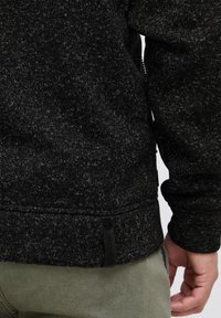 Black textured sweater with a speckled design, featuring a tag at the bottom and a zippered side. Worn over olive green pants.