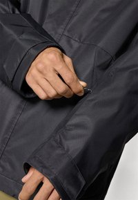 Main ajustant la fermeture éclair de la poche de la manche d'une veste noire avec des détails visibles sur le poignet et la couture.