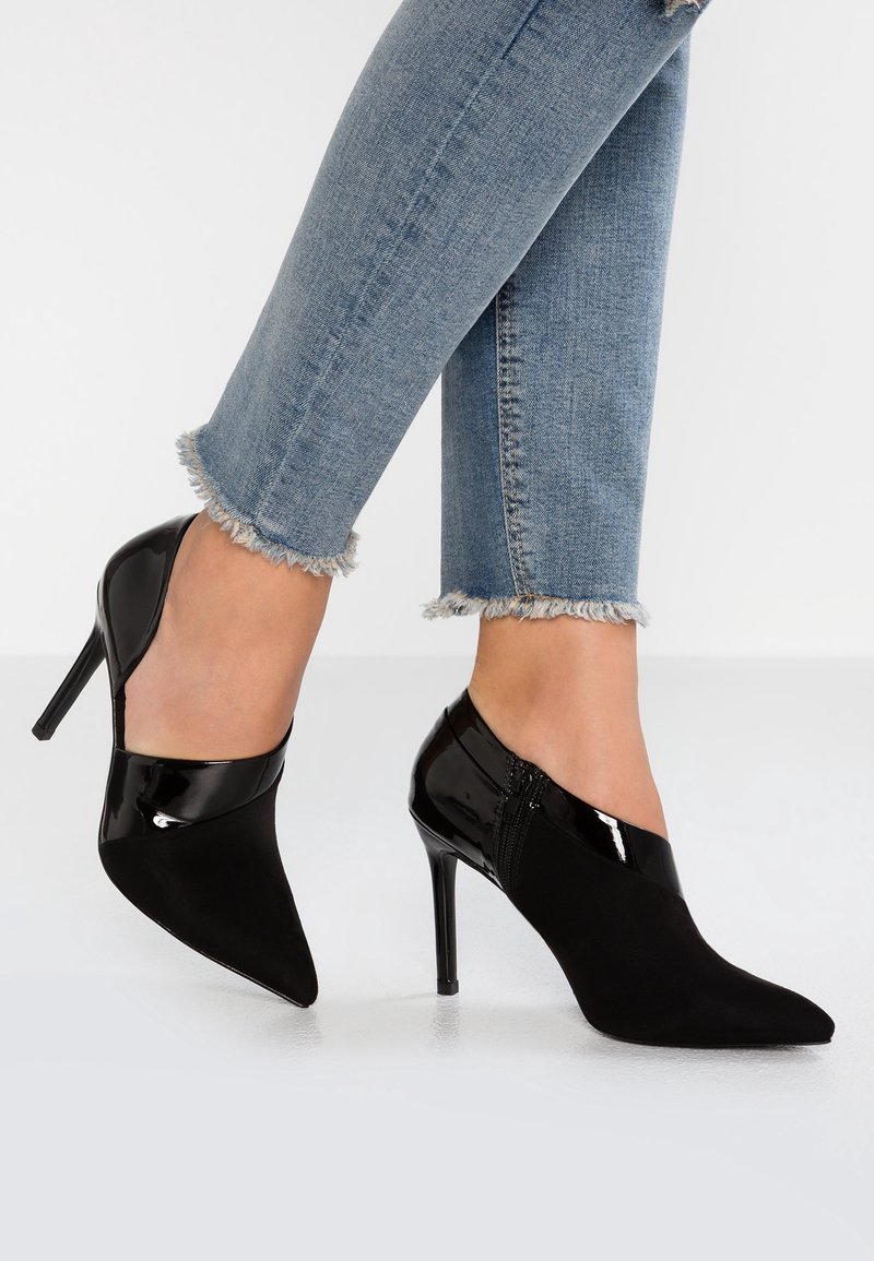 Bottines à talons aiguilles pointues noires avec détails en cuir verni, portées avec un jean bleu clair à ourlet effiloché sur fond blanc.
